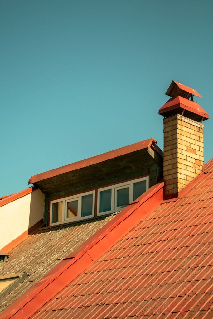 Red tiled house roof with a brick chimney and dormer window under a blue sky